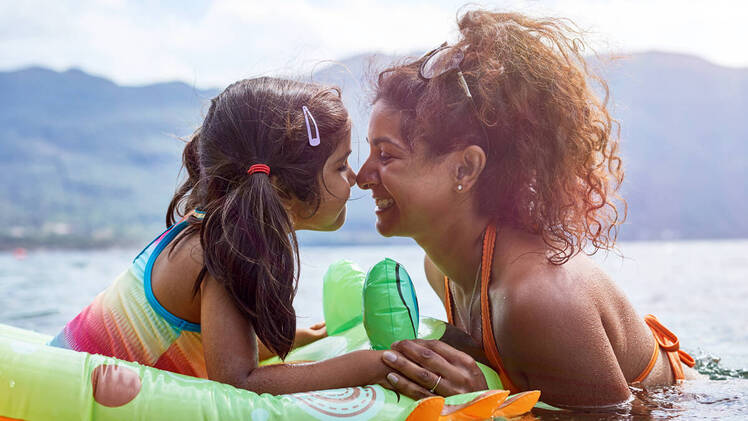 Mom with young daughter in a floatie swimming in a lake surrounded by nature