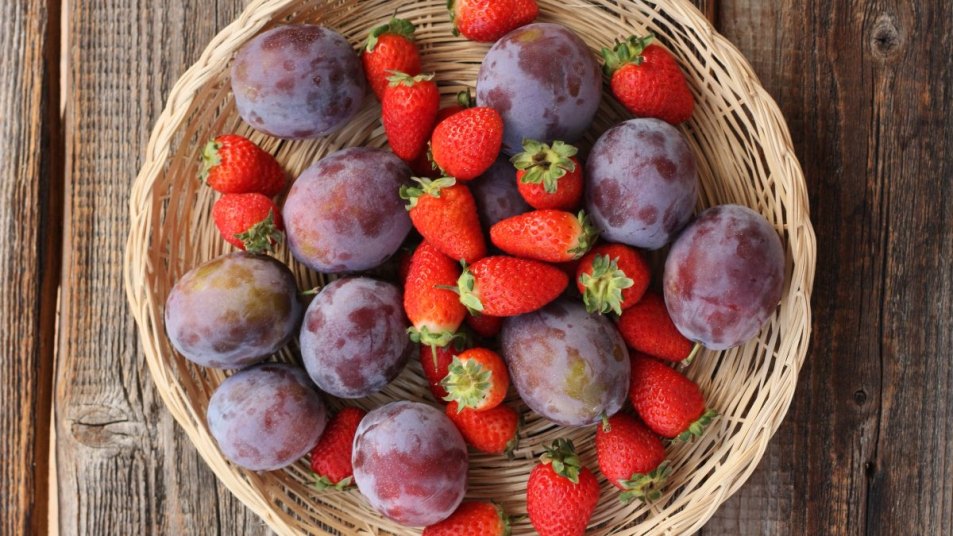 strawberries and plums in a woven basket on wood table, summer fruits