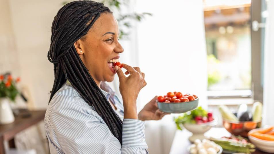 what to eat on ozempic: Photo of smiling Afro-American woman eating tomato in her kitchen during the day.