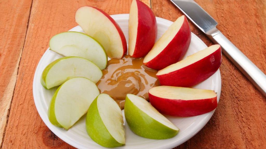 what to eat on ozempic: "Wedges of green and red apples surround a pool of organic peanut butter on a white plate, which rests on a rustic wood table."