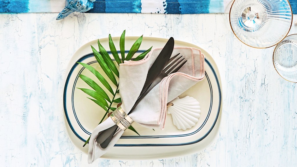 DIY beach decor: Contemporary nautical tablescape with a dinner plate topped with a palm leaf, seashell ornament and cutlery bundle alongside a blue and white stripy runner and gold-trimmed glassware 