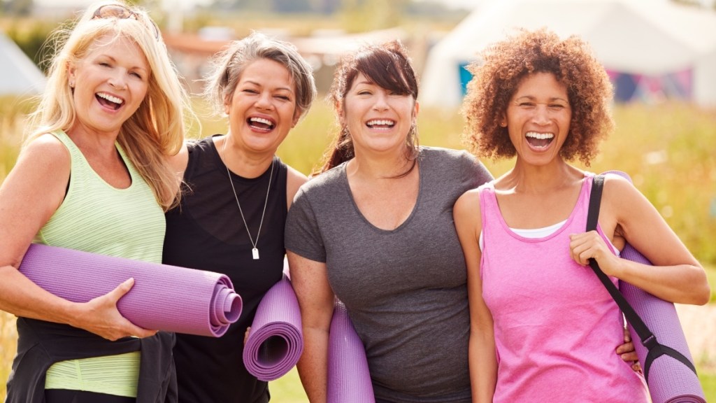 Four mature women holding yoga mats, smiling and laughing