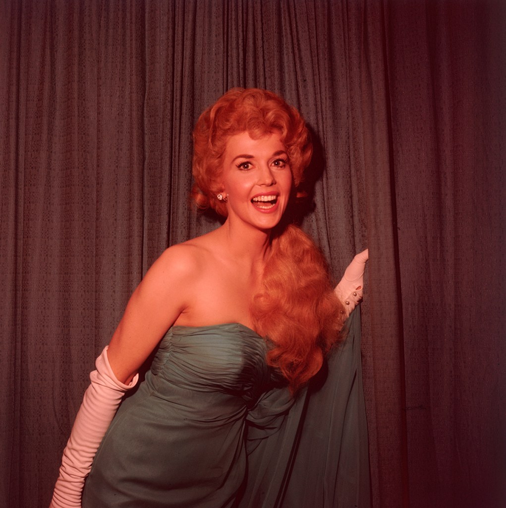 circa 1965:  Portrait of actor Donna Douglas, wearing a blue shoulderless gown and long white gloves, standing in front of a curtain and smiling