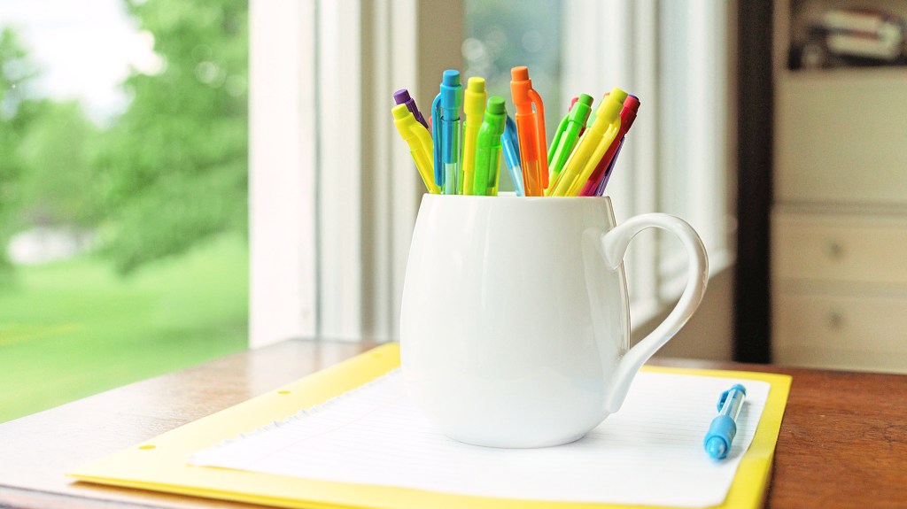 Home office organizing: Colorful pencils in a white mug set on top of a folder and notebook paper on a wooden desk