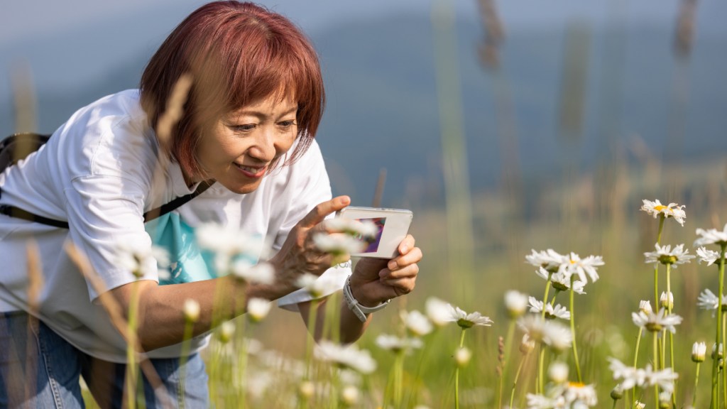 Older woman happily taking a close-up photo of a flower as she enjoys a flourishing life